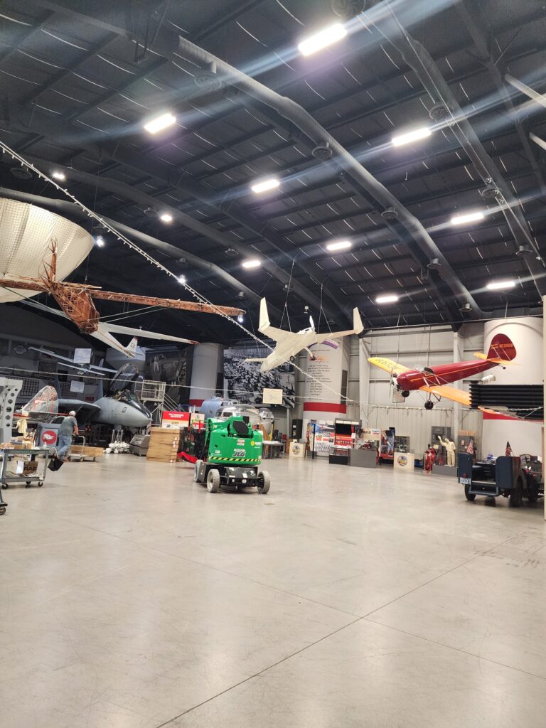 Interior of the Tulsa Air & Space Museum hangar with aircraft suspended from the ceiling, a green articulating manlift on the floor and rows of high-bay LED fixtures overhead