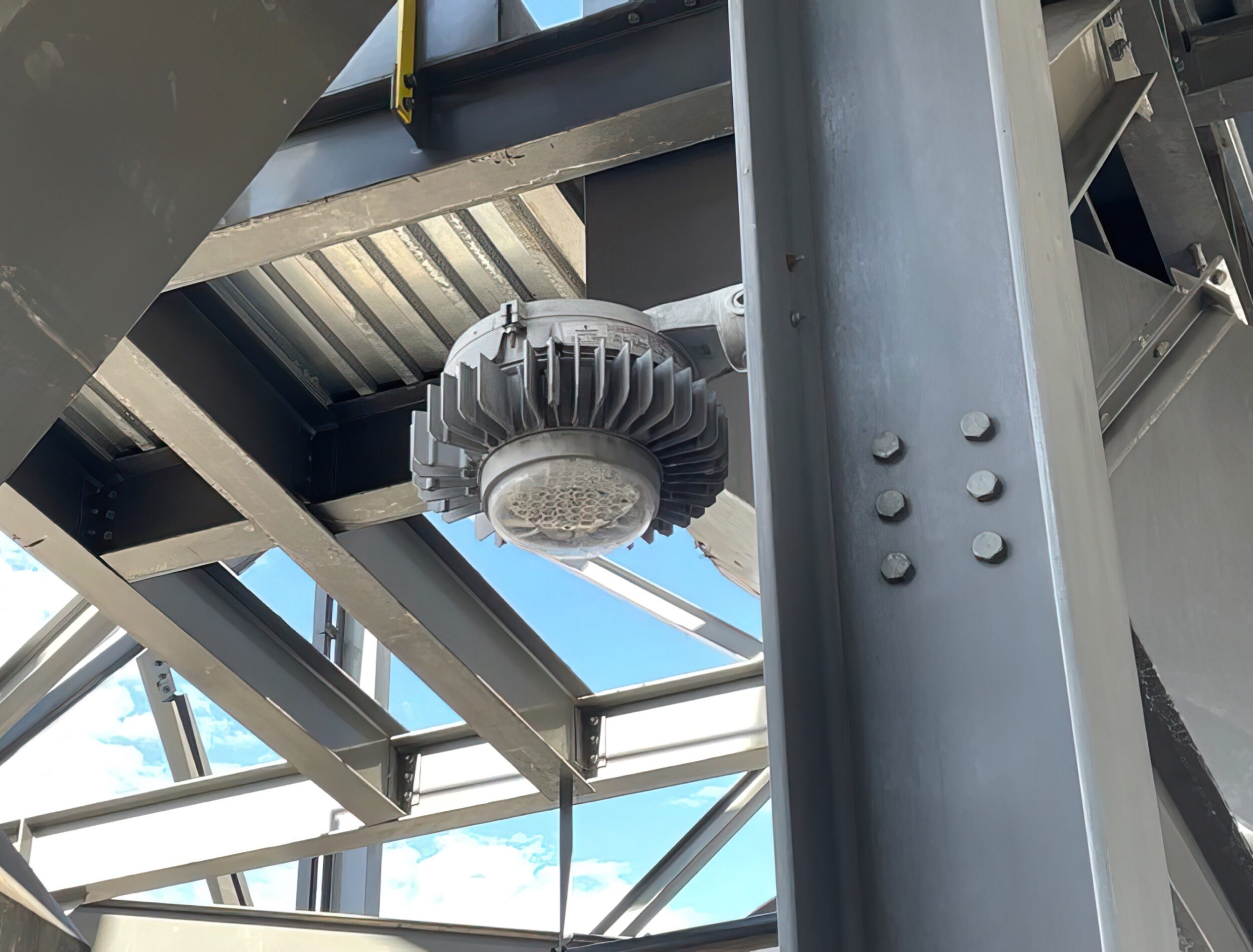 Appleton Mercmaster LED luminaire with a finned heat sink mounted under the steel framework of an industrial plant, blue sky visible through the structure