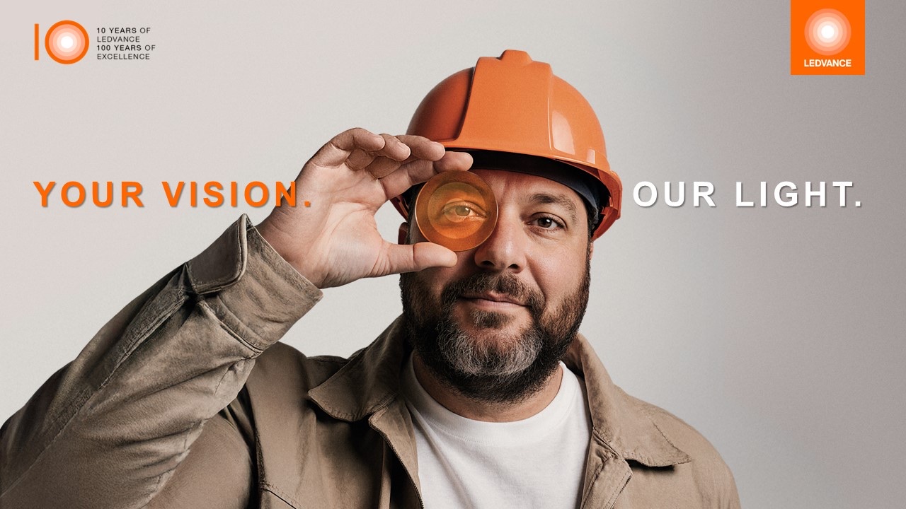 Bearded man in an orange hard hat holding an orange LEDVANCE disc over one eye, with the campaign line “Your Vision. Our Light.” and a 10-year anniversary mark