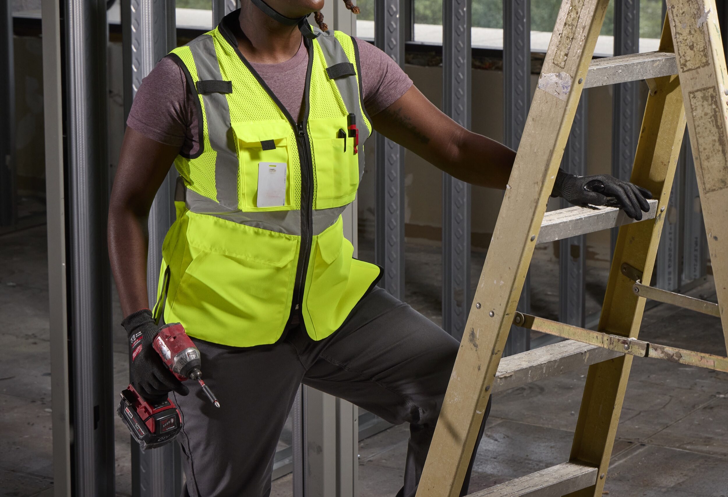 Woman on a jobsite wearing a Milwaukee yellow Class 2 high visibility safety vest with reflective stripes, standing beside a wooden ladder with a drill in hand